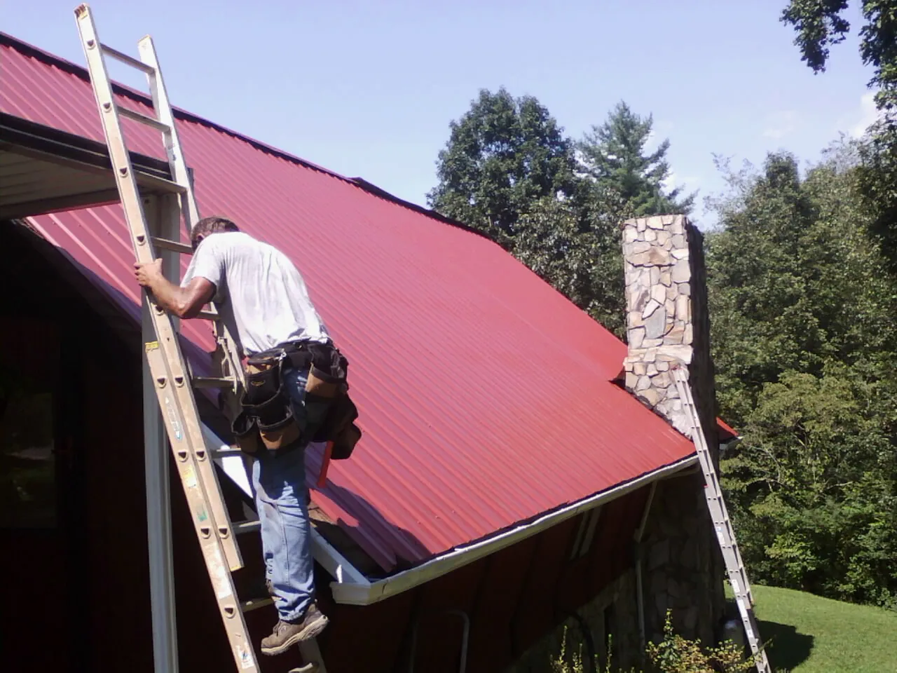 Roofer on a ladder beside a red metal roof and stone chimney — Soffit & Fascia Repair by Roofing Brothers in Boiceville